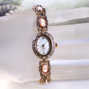 May include: A gold-toned vintage wristwatch with a white face and a decorative bezel set with small crystals. The watch band features cameo-style portraits and ornate detailing. The watch is an elegant accessory.