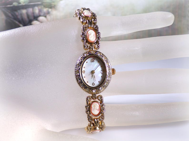May include: A gold-toned vintage wristwatch with a white face and a decorative bezel set with small crystals. The watch band features cameo-style portraits and ornate detailing. The watch is an elegant accessory.