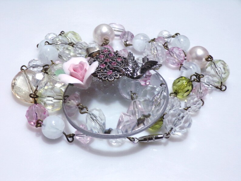 Peut inclure: Un collier d&eacute;licat avec un pendentif en verre transparent, orn&eacute; d'une fleur rose et d'un charme d'oiseau argent&eacute;. Le collier est compos&eacute; de perles de verre transparentes, roses et vertes.