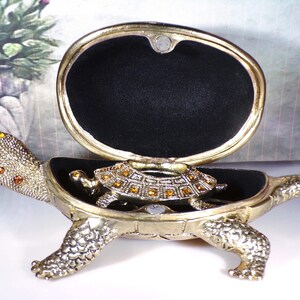 May include: A gold turtle trinket box with a hinged lid and a smaller turtle inside. The turtle is decorated with rhinestones.