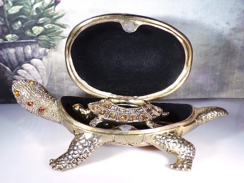 May include: A gold turtle trinket box with a hinged lid and a smaller turtle inside. The turtle is decorated with rhinestones.