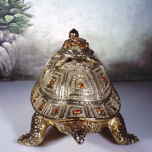 May include: A gold turtle trinket box with a detailed shell design and orange rhinestones. The turtle is sitting on a white surface.