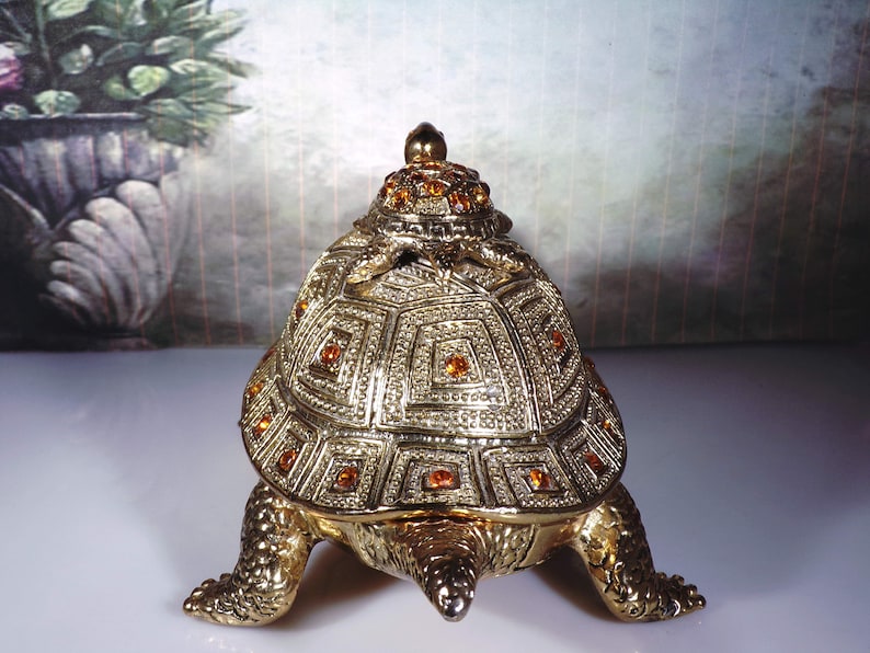 May include: A gold turtle trinket box with a detailed shell design and orange rhinestones. The turtle is sitting on a white surface.