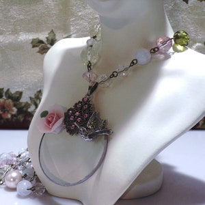 Peut inclure: Un collier d&eacute;licat avec un pendentif en forme de loupe orn&eacute; d'une fleur rose et d'un motif d'oiseau. Le collier est compos&eacute; de perles de verre transparentes, roses et vertes.