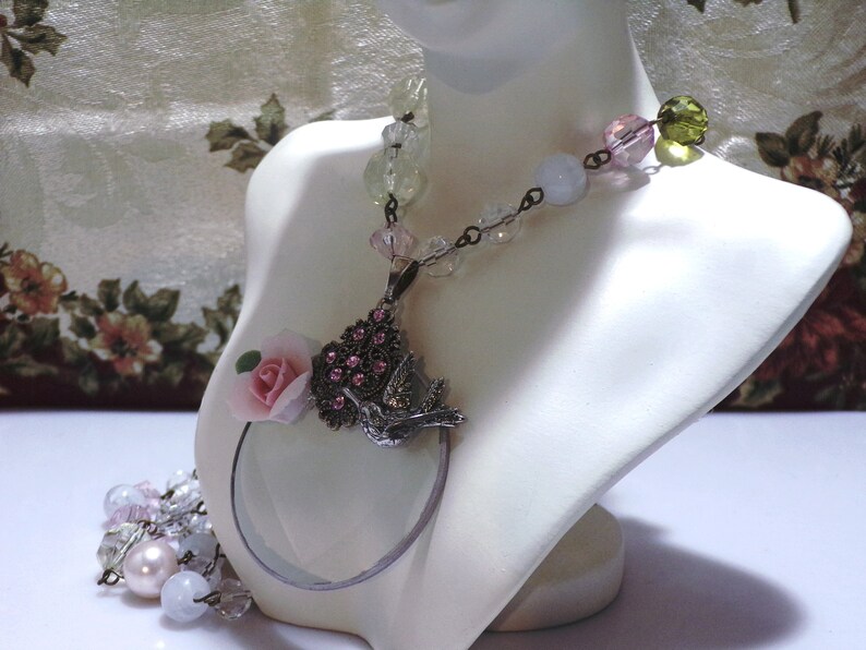 Peut inclure: Un collier d&eacute;licat avec un pendentif en forme de loupe orn&eacute; d'une fleur rose et d'un motif d'oiseau. Le collier est compos&eacute; de perles de verre transparentes, roses et vertes.