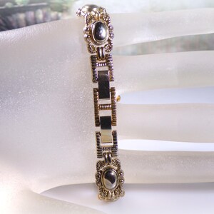 May include: A gold-toned bracelet with an oval, dark-colored stone at each end. The bracelet features rectangular and decorative links. The design includes floral and geometric patterns, creating an antique or vintage aesthetic.