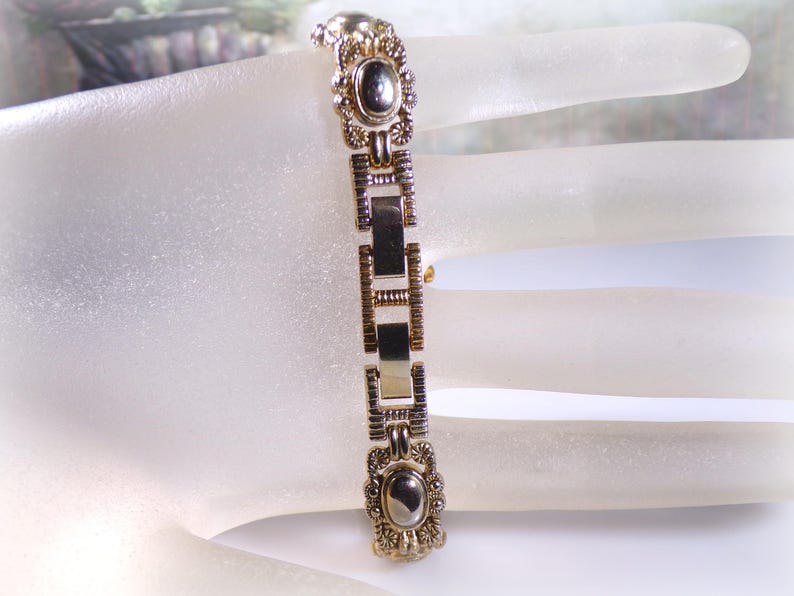 May include: A gold-toned bracelet with an oval, dark-colored stone at each end. The bracelet features rectangular and decorative links. The design includes floral and geometric patterns, creating an antique or vintage aesthetic.