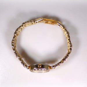 May include: A gold-toned wristwatch with a decorative band and a round face. The watch band is made of linked, ornate segments. The watch face is surrounded by small, sparkling stones. The watch is set against a white background.