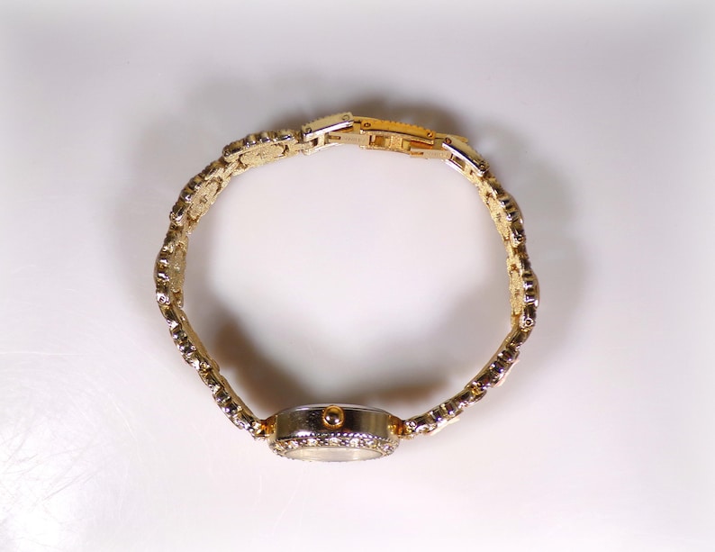 May include: A gold-toned wristwatch with a decorative band and a round face. The watch band is made of linked, ornate segments. The watch face is surrounded by small, sparkling stones. The watch is set against a white background.