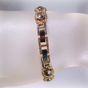 May include: A gold and silver-toned bracelet with a rectangular link design. The bracelet features decorative circular accents with a central silver-colored stone. The bracelet is displayed on a light-colored surface.