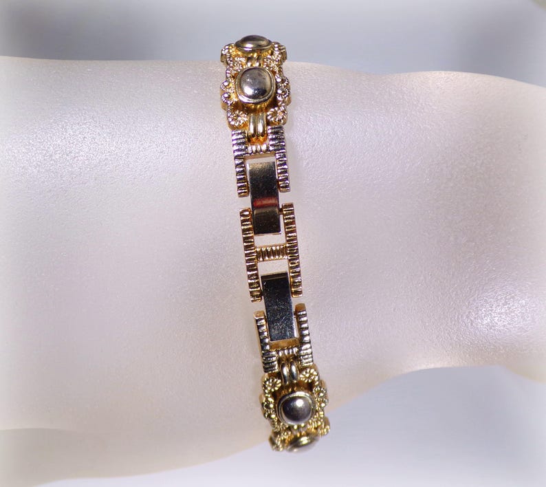 May include: A gold and silver-toned bracelet with a rectangular link design. The bracelet features decorative circular accents with a central silver-colored stone. The bracelet is displayed on a light-colored surface.