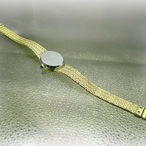 May include: A gold-coloured wristwatch featuring a woven strap and a round, silver watch face. The watch band has a mesh design, and the clasp is visible. The watch is shown on a light-coloured surface.