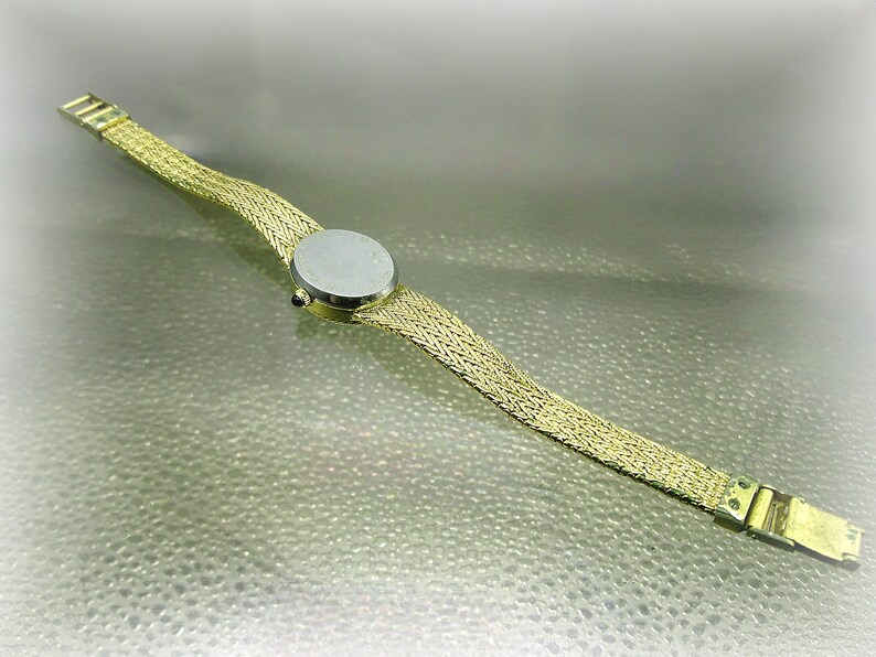 May include: A gold-coloured wristwatch featuring a woven strap and a round, silver watch face. The watch band has a mesh design, and the clasp is visible. The watch is shown on a light-coloured surface.