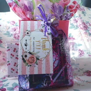 May include: A gift bag with a pink and white striped tag that says "Thank You for supporting my business." The bag is tied with a purple ribbon and contains a gift wrapped in floral tissue paper.