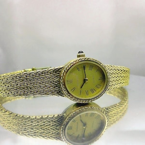 May include: A gold-coloured Movado quartz watch with a woven strap. The watch face is oval with Roman numerals and gold hands. The watch is reflected on a mirrored surface.