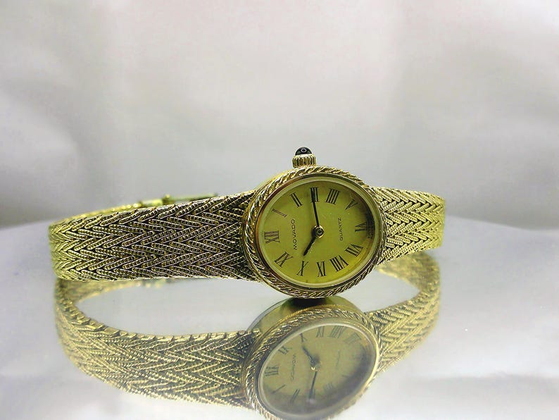 May include: A gold-coloured Movado quartz watch with a woven strap. The watch face is oval with Roman numerals and gold hands. The watch is reflected on a mirrored surface.