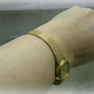 May include: A gold wristwatch with a woven strap is shown on a wrist. The watch face is round with a gold bezel. The strap has a textured, woven design. The watch is a classic accessory.