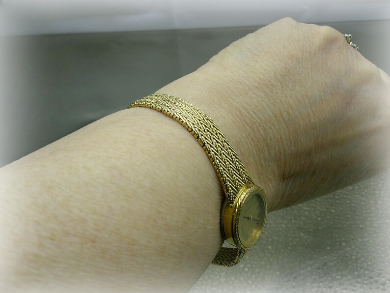 May include: A gold wristwatch with a woven strap is shown on a wrist. The watch face is round with a gold bezel. The strap has a textured, woven design. The watch is a classic accessory.