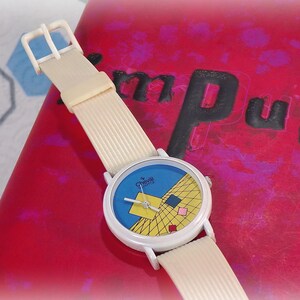 May include: A wristwatch with a cream-colored ribbed strap and a white round case. The watch face features a blue background with a yellow grid pattern, a large yellow square, and smaller red, blue, and pink squares. The text "Chevill Quartz" is printed on the face.