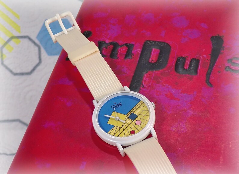 May include: A wristwatch with a cream-colored ribbed strap and a white round case. The watch face features a blue background with a yellow grid pattern, a large yellow square, and smaller red, blue, and pink squares. The text "Chevill Quartz" is printed on the face.