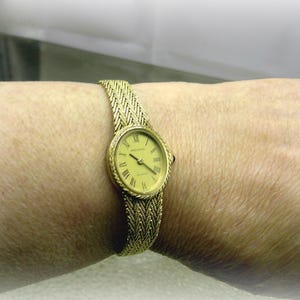 May include: A gold-coloured wristwatch with a woven strap is shown on a wrist. The watch face is oval with Roman numerals and hands indicating the time. The strap and watch casing have a matching gold colour.
