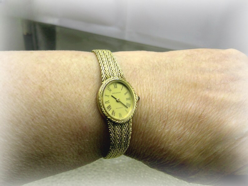 May include: A gold-coloured wristwatch with a woven strap is shown on a wrist. The watch face is oval with Roman numerals and hands indicating the time. The strap and watch casing have a matching gold colour.