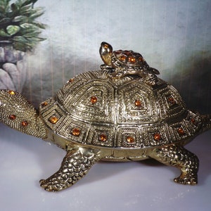 May include: A gold-colored turtle figurine with a smaller turtle on its back. Both turtles are decorated with orange rhinestones.