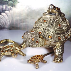 May include: Three gold-tone turtle figurines with amber-colored rhinestones. The largest turtle has a smaller turtle on its back.