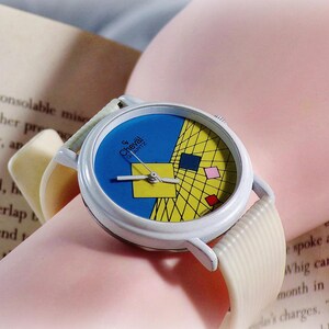 May include: A Cheval Quartz wristwatch with a white case and a textured off-white strap. The watch face features a blue and yellow geometric grid pattern with small red, pink, and yellow squares, along with silver-toned hour, minute, and second hands.