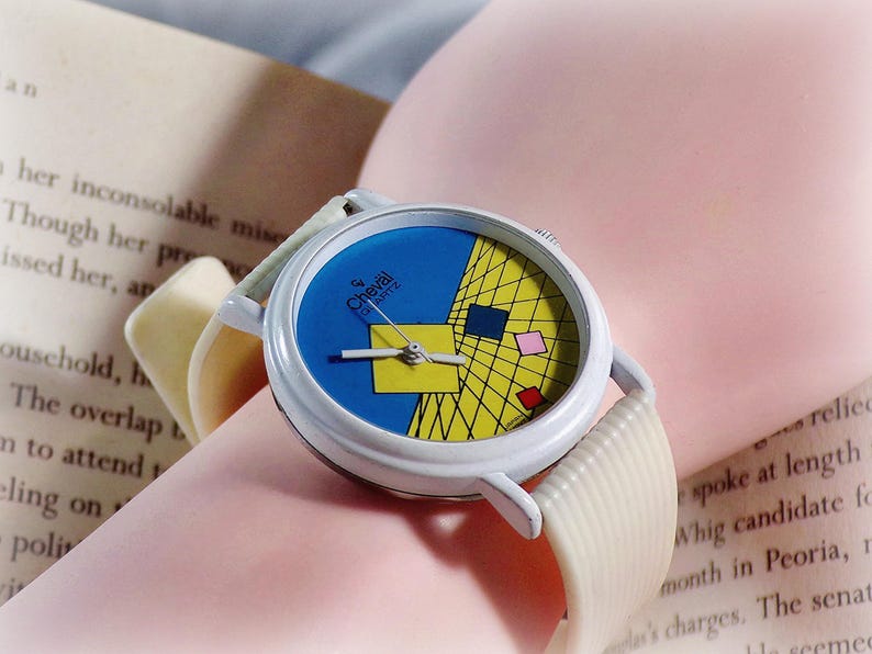 May include: A Cheval Quartz wristwatch with a white case and a textured off-white strap. The watch face features a blue and yellow geometric grid pattern with small red, pink, and yellow squares, along with silver-toned hour, minute, and second hands.