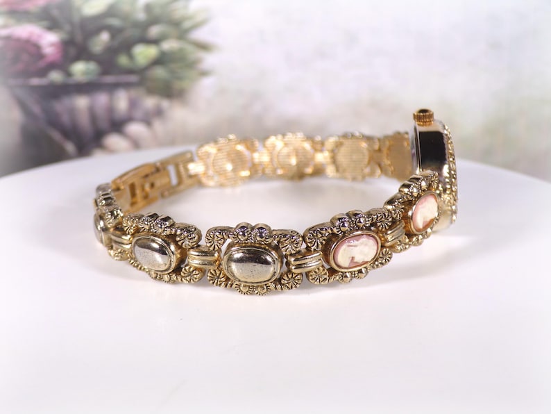 May include: Gold-toned bracelet watch with oval and cameo-style accents. The watch features a decorative link design with a clasp closure. The watch face is rectangular and has a gold-tone finish. The bracelet is approximately 7 inches in length.