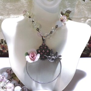 Peut inclure: Un collier en argent avec un pendentif en forme de loupe orn&eacute; d'une fleur en porcelaine rose et d'un colibri. Le collier est compos&eacute; de perles de verre transparentes et roses.