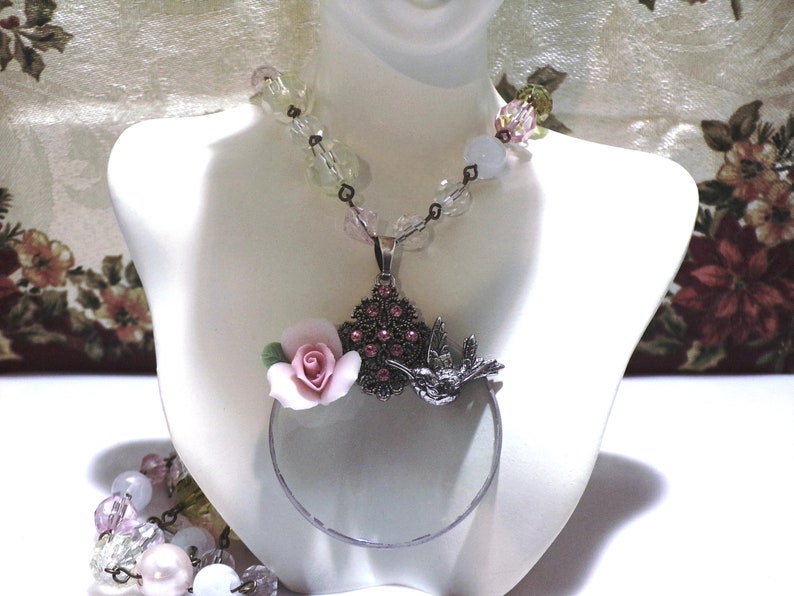 Peut inclure: Un collier en argent avec un pendentif en forme de loupe orn&eacute; d'une fleur en porcelaine rose et d'un colibri. Le collier est compos&eacute; de perles de verre transparentes et roses.