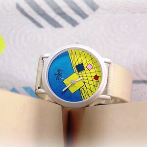 May include: A Cheval Quartz wristwatch with a white case and a textured off-white strap. The watch face features a blue and yellow geometric design with a grid pattern, small colored squares, and silver hands. The text Cheval Quartz is printed on the face.