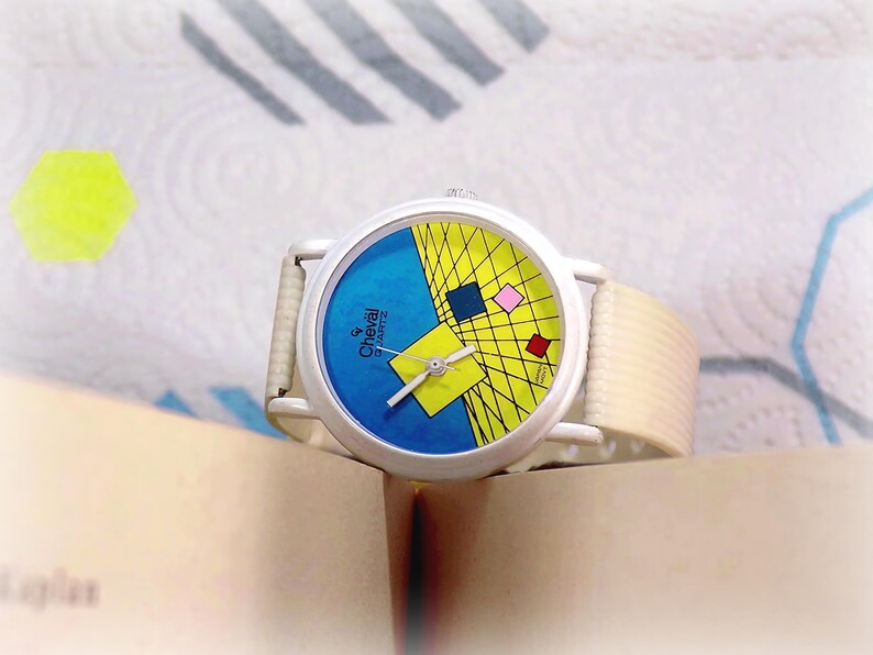 May include: A Cheval Quartz wristwatch with a white case and a textured off-white strap. The watch face features a blue and yellow geometric design with a grid pattern, small colored squares, and silver hands. The text Cheval Quartz is printed on the face.
