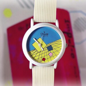May include: A white wristwatch with a ribbed white strap and a round white case. The watch face features a blue background with a yellow grid pattern, a large yellow square, and smaller blue, pink, and red squares. The text reads Cheval Quartz.