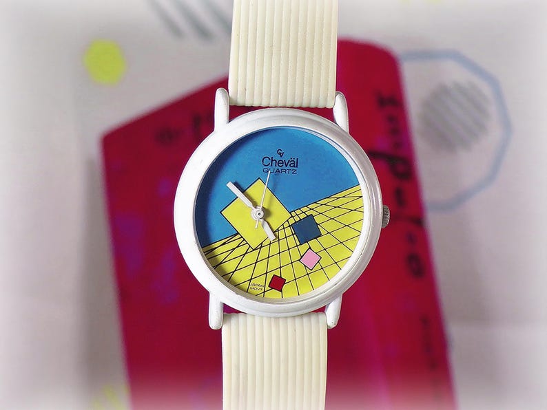 May include: A white wristwatch with a ribbed white strap and a round white case. The watch face features a blue background with a yellow grid pattern, a large yellow square, and smaller blue, pink, and red squares. The text reads Cheval Quartz.