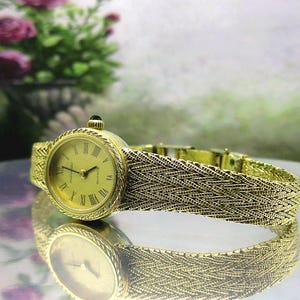 May include: A gold-coloured wristwatch with a woven strap and a round face with Roman numerals. The watch has a small, dark-coloured stone on the crown. The watch is displayed on a reflective surface.