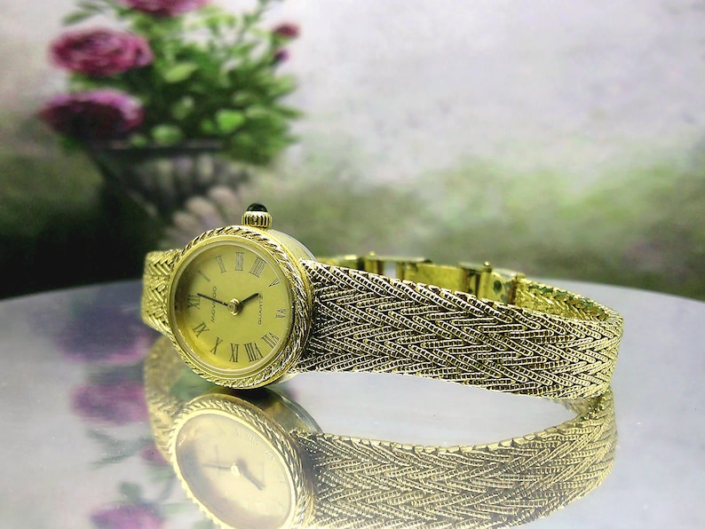 May include: A gold-coloured wristwatch with a woven strap and a round face with Roman numerals. The watch has a small, dark-coloured stone on the crown. The watch is displayed on a reflective surface.