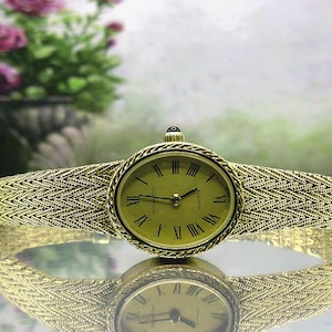 May include: A gold-coloured Movado quartz watch with a woven strap. The oval watch face features black Roman numerals and hands. The strap has a textured, herringbone pattern. The watch is reflected on a shiny surface.