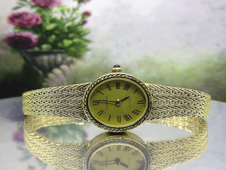 May include: A gold-coloured Movado quartz watch with a woven strap. The oval watch face features black Roman numerals and hands. The strap has a textured, herringbone pattern. The watch is reflected on a shiny surface.