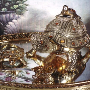 May include: Two gold-toned turtle figurines with jeweled accents. The larger turtle is on a tray with a mirror surface. The smaller turtle is on the tray and reflected in the mirror.