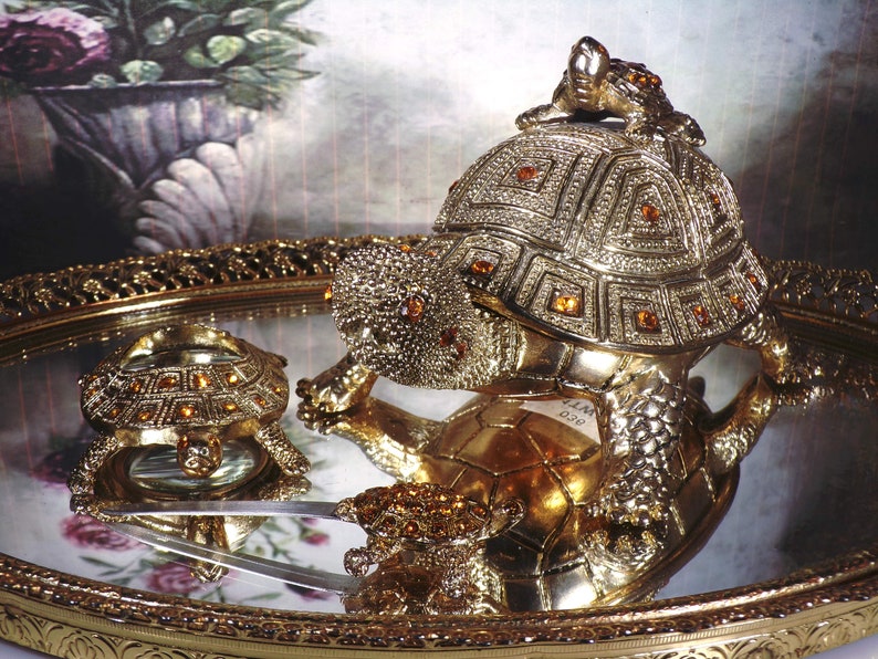 May include: Two gold-toned turtle figurines with jeweled accents. The larger turtle is on a tray with a mirror surface. The smaller turtle is on the tray and reflected in the mirror.