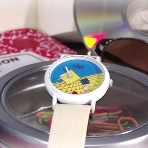 May include: A Cheval Quartz wristwatch with a white case and a textured off-white strap. The watch face features a blue upper section and a yellow lower section with a black grid pattern, accented by small floating squares in yellow, blue, and pink.