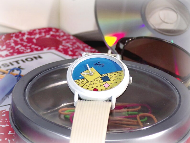 May include: A Cheval Quartz wristwatch with a white case and a textured off-white strap. The watch face features a blue upper section and a yellow lower section with a black grid pattern, accented by small floating squares in yellow, blue, and pink.