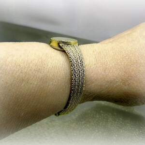 May include: A gold-coloured bracelet featuring a woven band and a circular centrepiece, possibly with a stone. The bracelet is worn on a wrist, highlighting its texture and design. The band is approximately 2 cm wide.