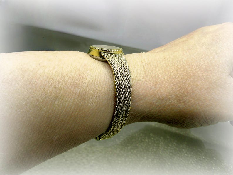 May include: A gold-coloured bracelet featuring a woven band and a circular centrepiece, possibly with a stone. The bracelet is worn on a wrist, highlighting its texture and design. The band is approximately 2 cm wide.