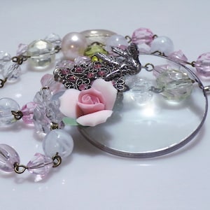 Peut inclure: Un collier d&eacute;licat avec un pendentif loupe, orn&eacute; d'une rose rose, de perles de verre transparentes et vertes, et d'un charme d'oiseau argent&eacute; avec des strass roses.