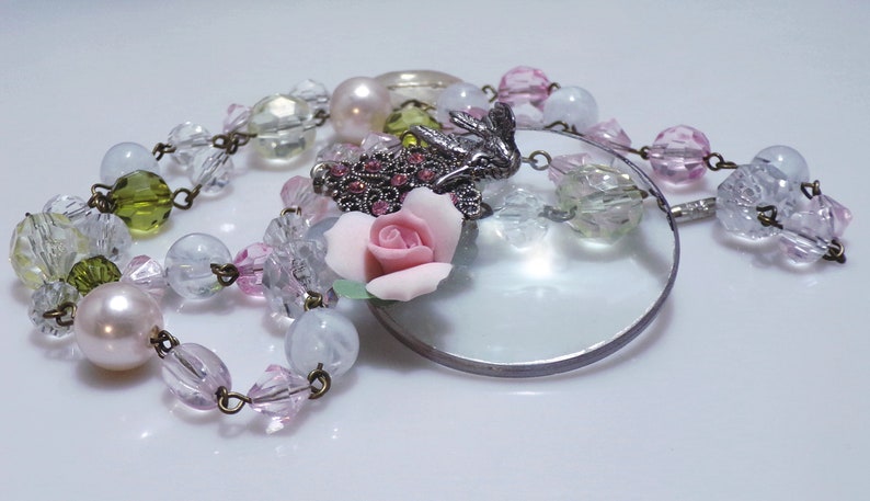 Peut inclure: Un collier d&eacute;licat avec un pendentif loupe, orn&eacute; d'une rose rose, de perles de verre transparentes et vertes, et d'un charme d'oiseau argent&eacute; avec des strass roses.