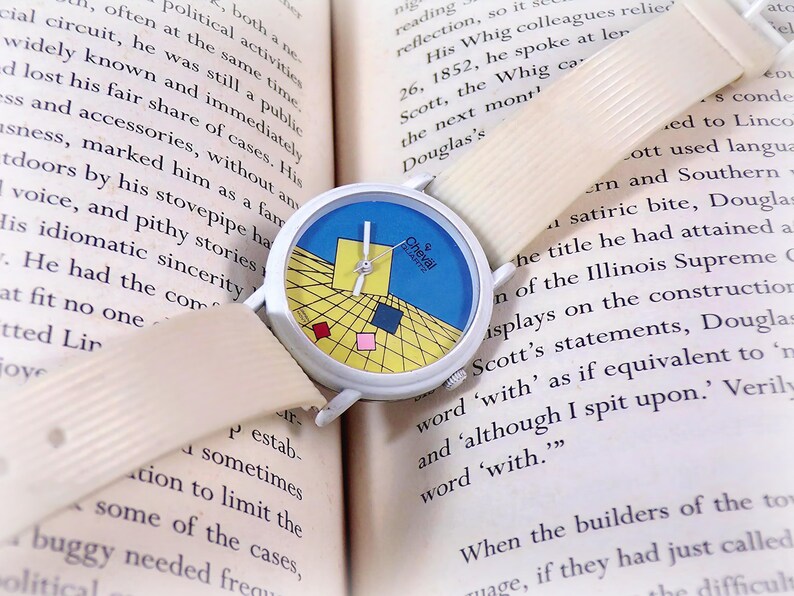 May include: A Cheval Quartz wristwatch with a white case and a textured off-white strap. The watch face features a blue and yellow grid pattern with small red, pink, and blue squares, white hands, and the text Cheval Quartz printed on the dial.